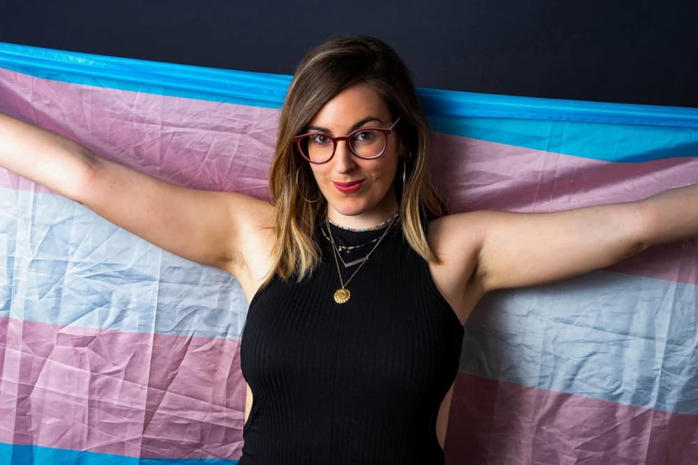 Mulher trans sorrindo e olhando para a câmera, com a bandeira trans ao fundo